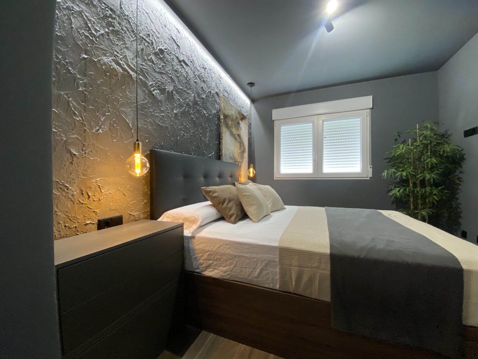 Dormitorio reformado con cabecero integrado, pared texturizada, apliques colgantes y ventana con persiana.