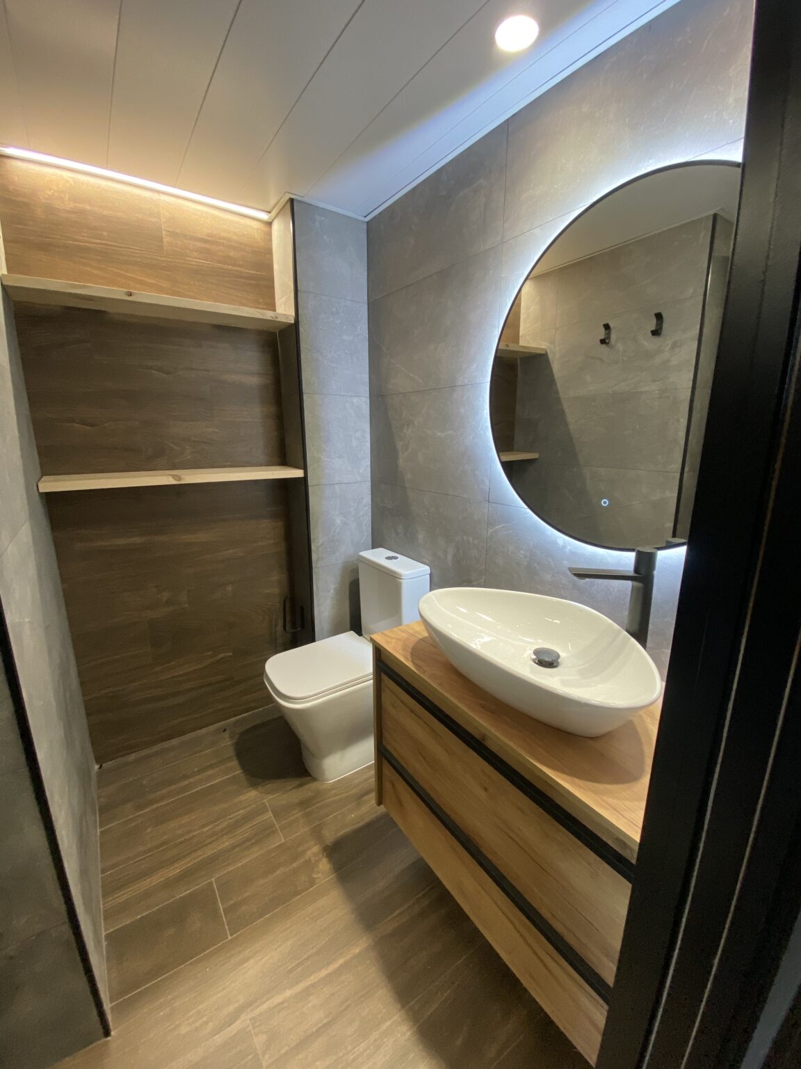 Aseo moderno con lavabo sobre mueble de madera, espejo circular retroiluminado y revestimiento porcelánico.