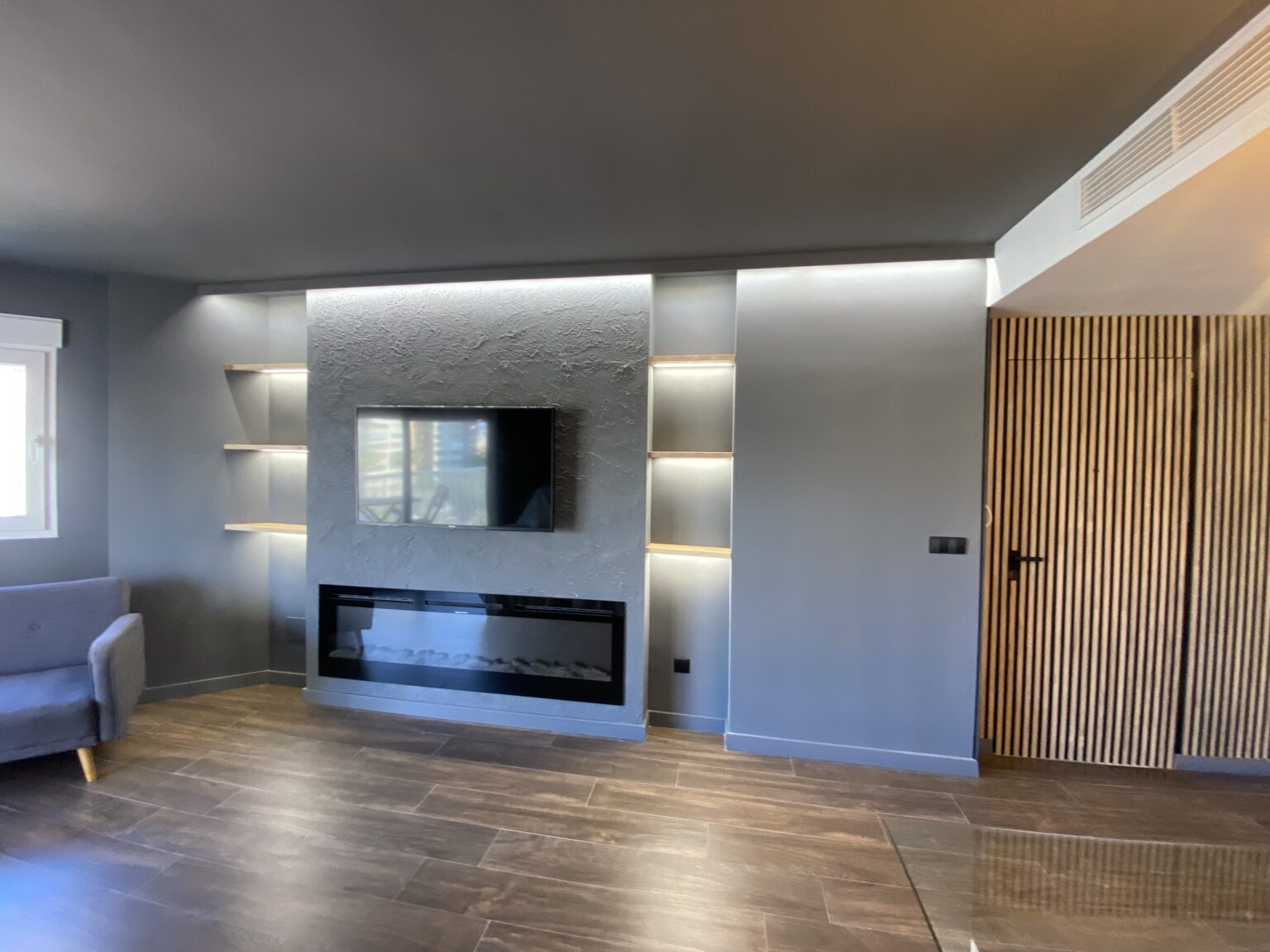 Salón reformado con mueble TV empotrado, estanterías e iluminación indirecta LED y suelo laminado.